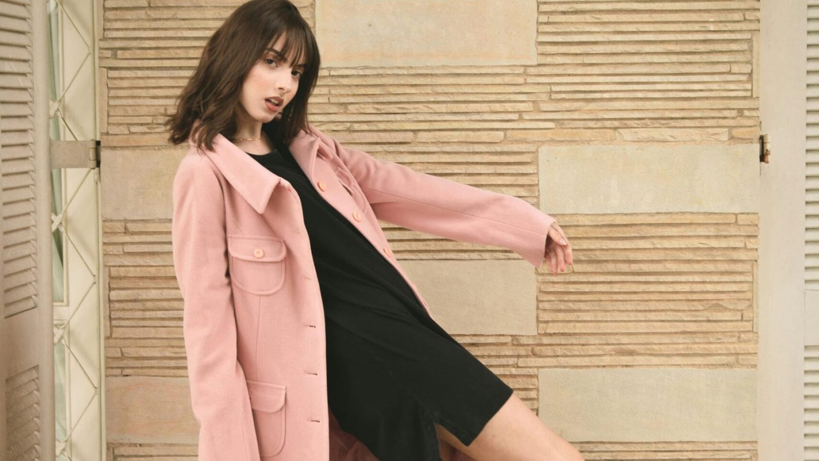 Fashion model in pink coat and black dress striking a dynamic pose indoors with stylish black heels.